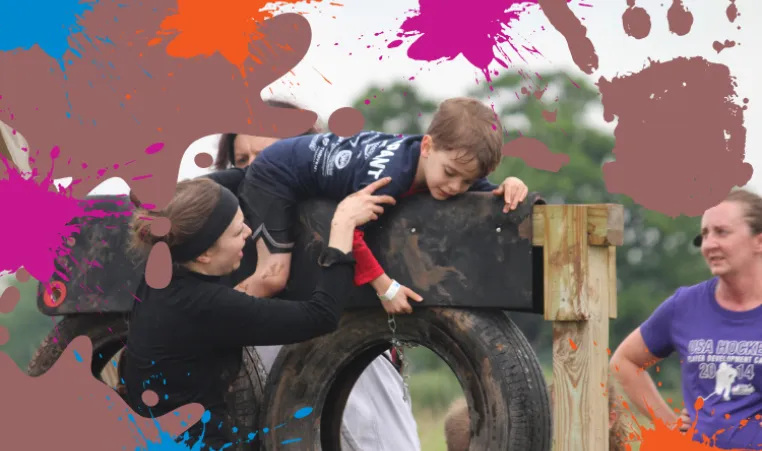 A photo of a few volunteers helping a child over a muddy tire obstacle during the mud run. 