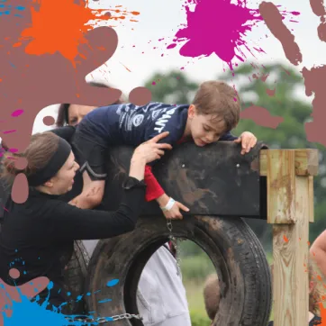 A photo of a few volunteers helping a child over a muddy tire obstacle during the mud run. 