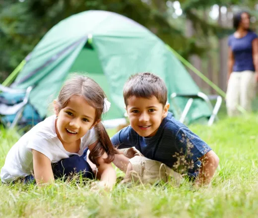 two children playing in the grass in front of their tent while a parent supervises.
